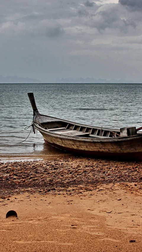 Ein langes, verwittertes Holzboot mit hochgezogenem Bug liegt auf einem steinigen Sandstrand am Meer, im Hintergrund ein bewölkter grauer Himmel.