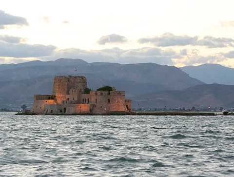 Eine steinerne Festung mit Rundturm auf einer kleinen Insel im Meer, im warmen Abendlicht, dahinter Bergketten unter bewölktem Himmel.
