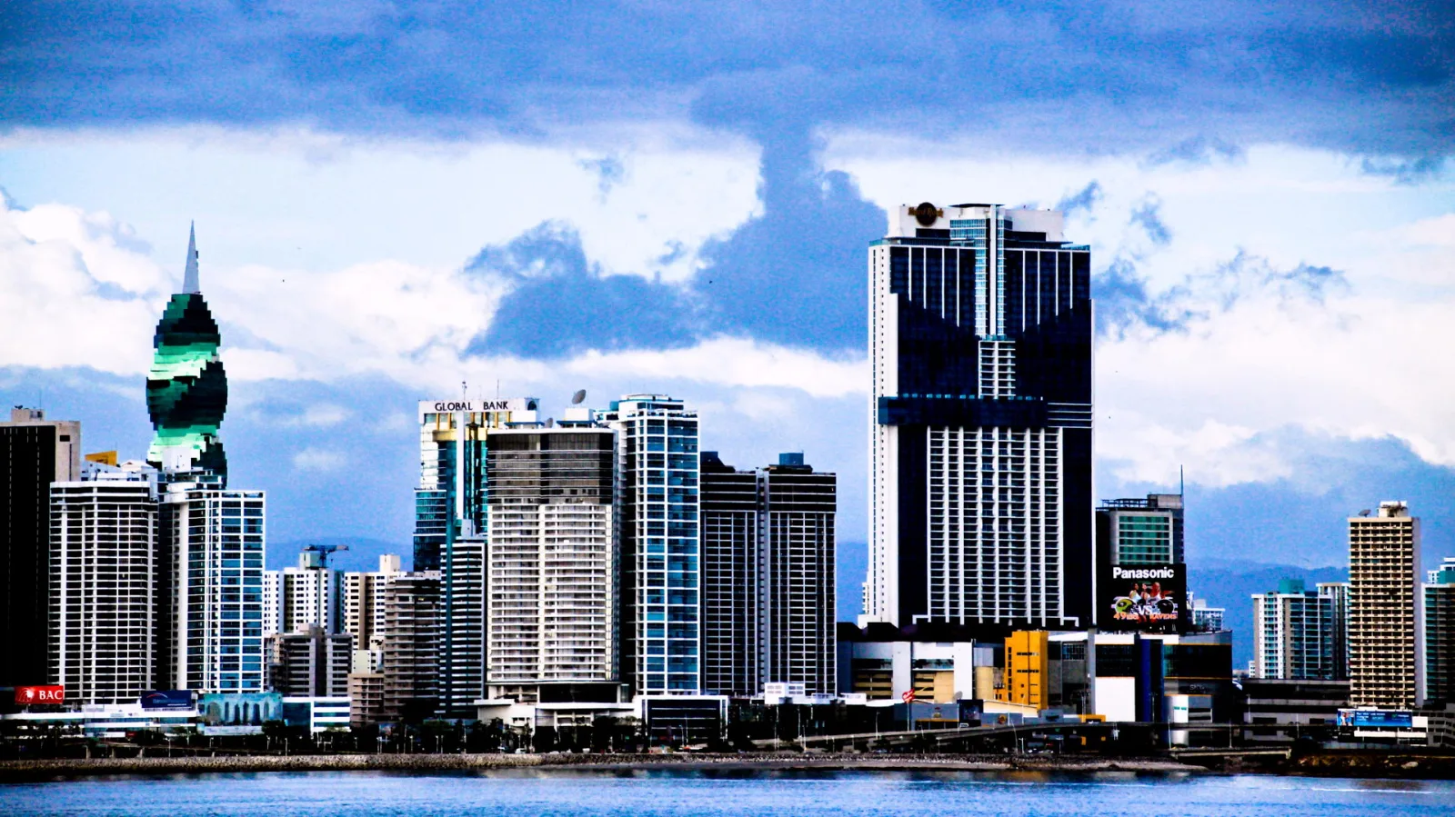 Skyline mit mehreren Hochhäusern hinter einer Uferpromenade, darüber eine bewölkte Himmelsfläche. Auffällig sind ein hoher dunkler Turm in der Bildmitte und ein Turm mit grün gestreifter Fassade links. Im Vordergrund liegt das Wasser.