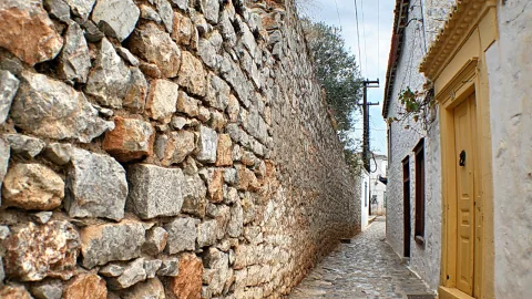Eine schmale Gasse mit hoher Bruchsteinmauer links, Kopfsteinpflaster im Vordergrund und einer gelben Holztür an einem weiß getünchten Haus rechts. Im Hintergrund ragt ein Olivenbaum über die Mauer, darüber hängen Stromleitungen am hellen Himmel.