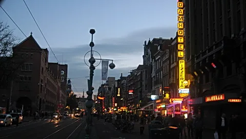 Abendliche Straße in Amsterdam mit Leuchtreklamen, darunter ein vertikales Schild Dam Hotel und eine Pizzeria, links ein historisches Backsteingebäude und eine Laterne.