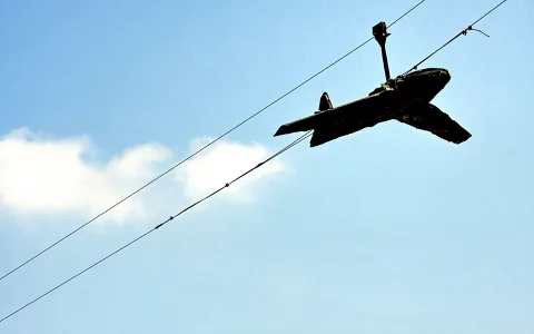 Dunkles Spielzeugflugzeug hängt an zwei Drähten vor blauem Himmel, links eine kleine weiße Wolke.