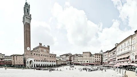 Weiter, heller Platz mit hohem Backsteinturm und mittelalterlichem Palast links, Menschen auf dem Pflaster und rechts Cafés mit roten Sonnenschirmen unter bewölktem Himmel.
