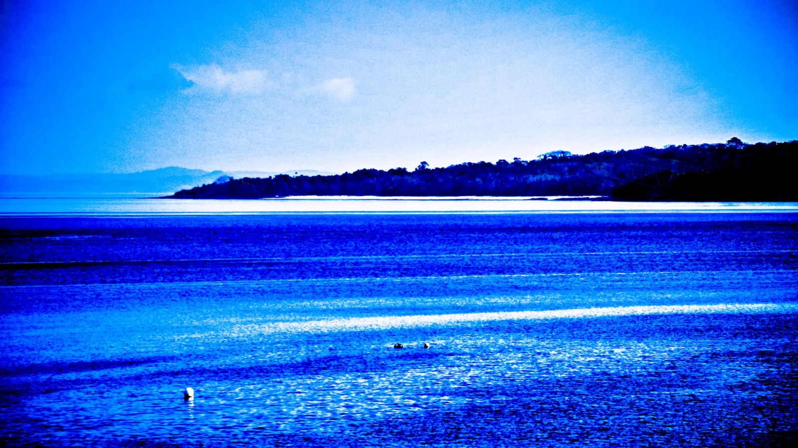 Weite blaue Wasserfläche mit glitzernden Reflexionen, im Hintergrund eine dunkel bewaldete Insel unter hellblauem Himmel, im Vordergrund zwei kleine Bojen oder Schwimmkörper.