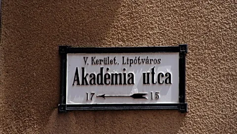 Weißes Straßenschild mit schwarzer Schrift „Akadémia utca“ und Angabe „V. Kerület, Lipótváros“ an einer braunen, strukturierten Hauswand, darunter die Hausnummern 15 und 17 mit Pfeil.
