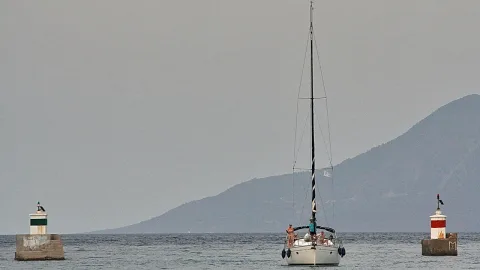 Ein weißes Segelboot mit hohem Mast fährt auf ruhigem, graublauem Meer auf eine Hafeneinfahrt zu, links und rechts stehen kleine Molenfeuer, im Hintergrund zeigt sich eine dunstige Insel.