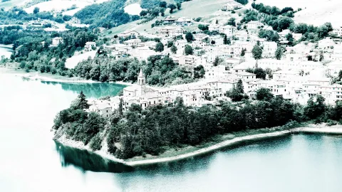 Hangdorf mit hellen Häusern und einem Kirchturm über einem See, im Vordergrund eine bewaldete Landzunge, blaugrün getönt