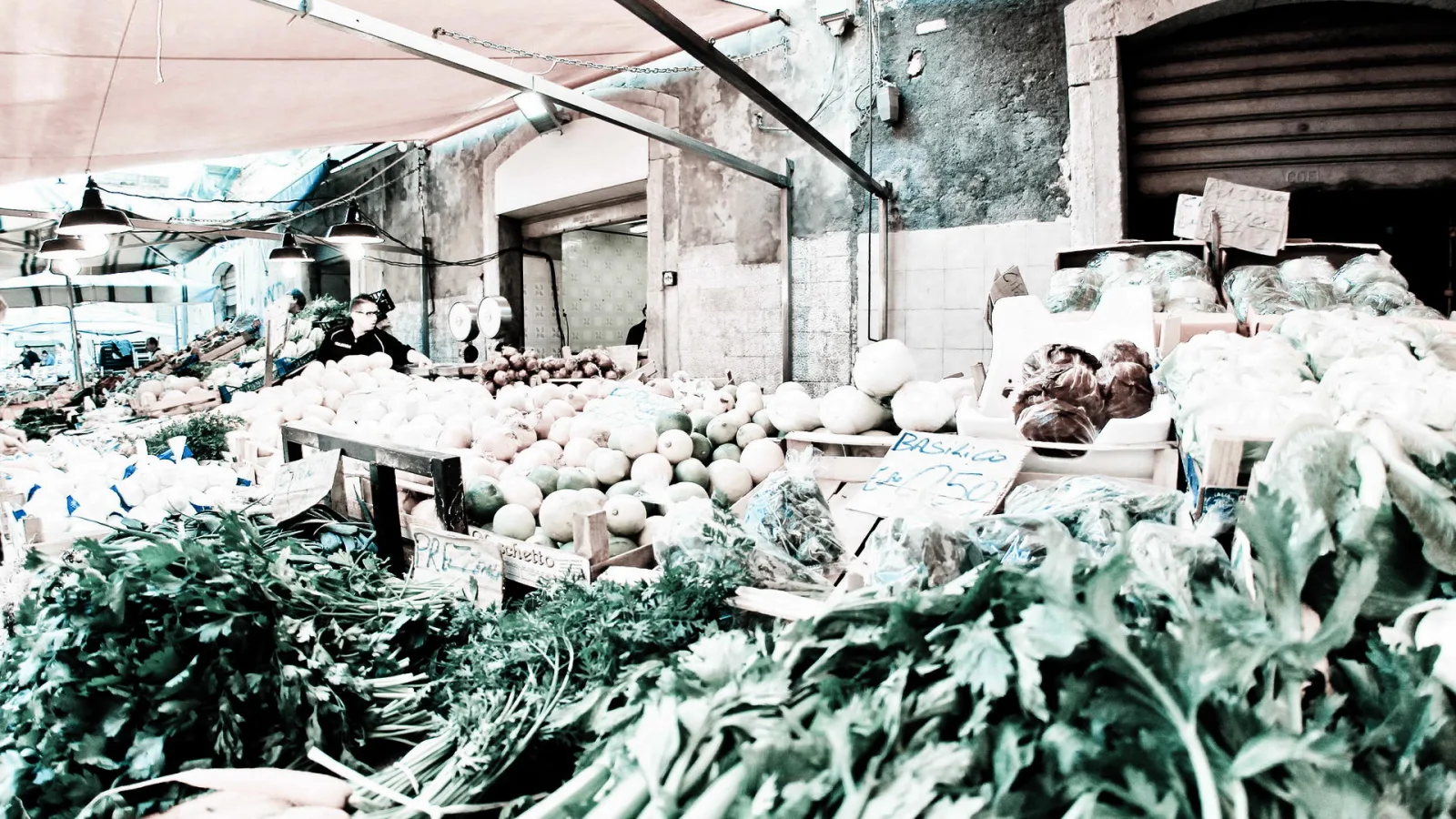 Gemüsestand auf einem Markt mit Fenchel, Blattgemüse und hellen Kürbissen, im Hintergrund Verkäufer unter einer Markise und alte Hausfassade
