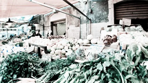 Gemüsestand auf einem Markt mit Fenchel, Blattgemüse und hellen Kürbissen, im Hintergrund Verkäufer unter einer Markise und alte Hausfassade
