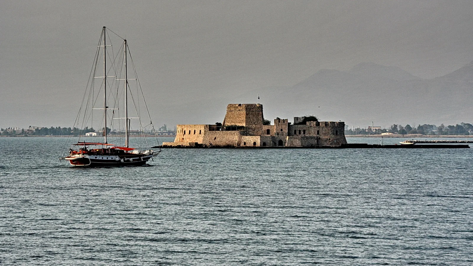 Steinerne Festung auf einer kleinen Insel im Meer, links ein Segelboot mit rotem Streifen, dahinter verschwommene Berge unter grauem Himmel.