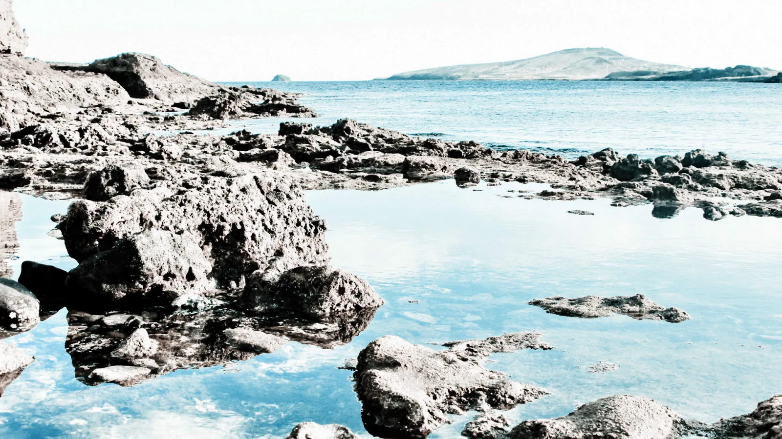 Dunkle, zerklüftete Felsen ragen aus flachem, türkisblauem Wasser. Im Hintergrund liegt eine flache Insel unter hellem Himmel.