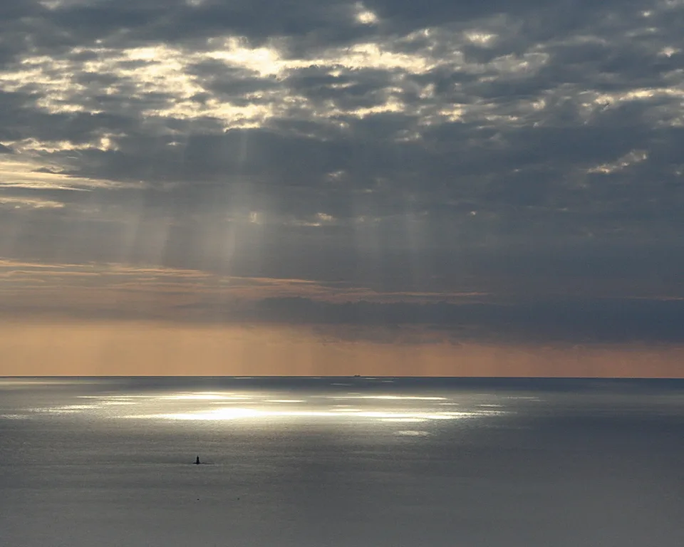 Sonnenstrahlen brechen durch Wolken auf eine glatte Meeresfläche, im Vordergrund ein kleiner dunkler Gegenstand auf dem Wasser, am Horizont ein Schiff.