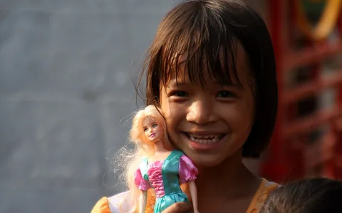 Lachendes Mädchen mit Pagenkopf hält eine blonde Puppe im bunten Kleid vor die Schulter, im Hintergrund eine graue Wand und rote Gitter.