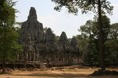 Der Bayon-Tempel mit mehreren hohen Steintürmen und Gesichtern, davor ein staubiger Vorplatz und Bäume am Rand.