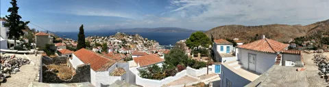 Panoramablick auf ein dicht bebautes Küstendorf mit weißen Häusern und roten Ziegeldächern, im Hintergrund das blaue Meer und eine Bergkette unter blauem Himmel mit Wolken.