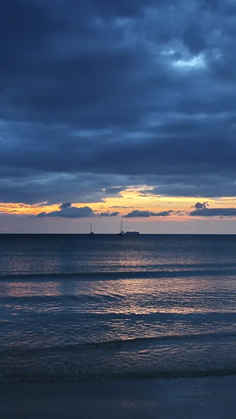 Meer in der Dämmerung unter dunklen Wolken, orangefarbener Horizont mit zwei Segelbooten, Wellen am Strand