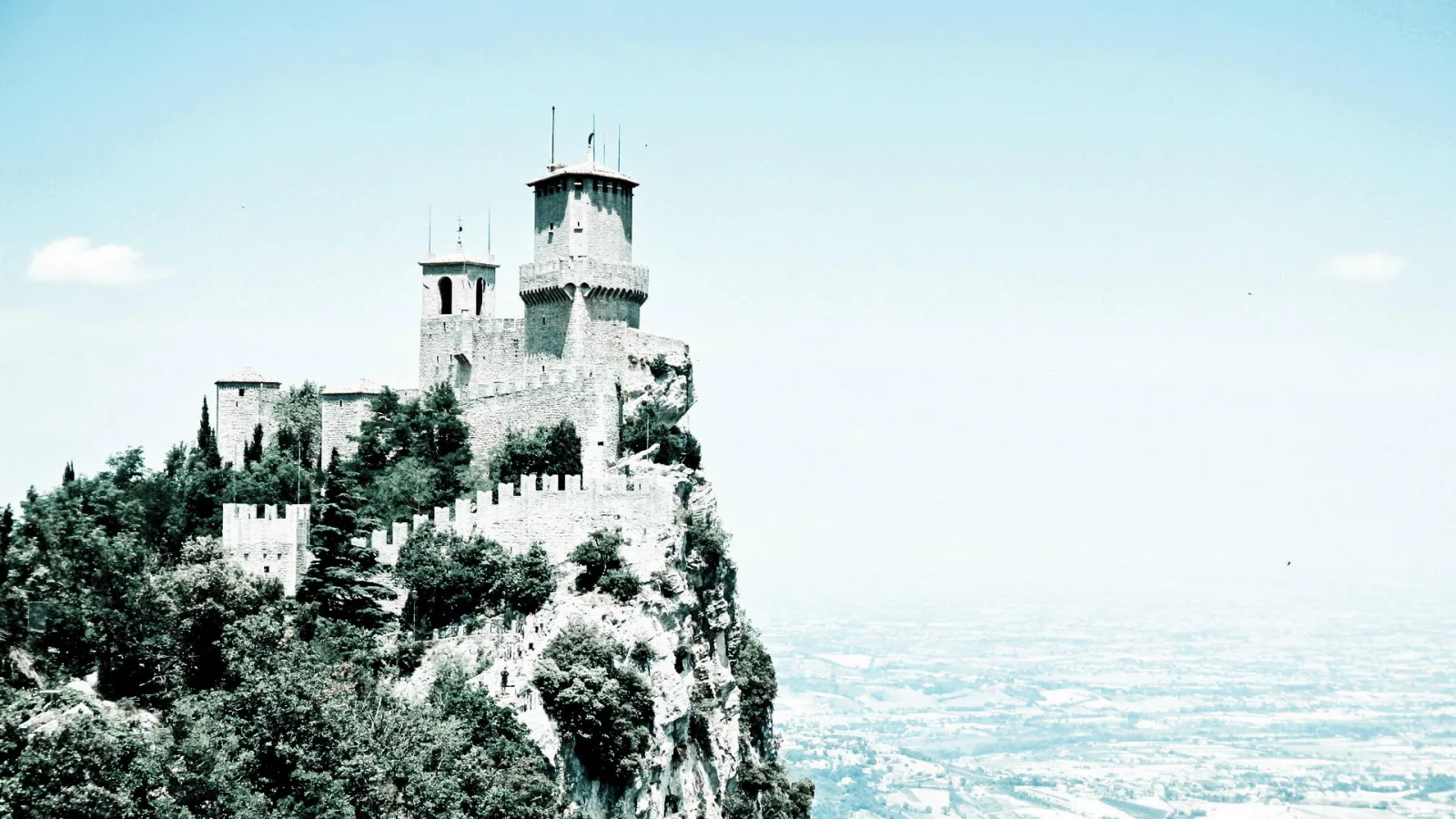 Mittelalterliche Festung mit Türmen und Zinnenmauern auf einem bewaldeten Felsen, im Hintergrund weite, hell diesige Ebene unter blassblauem Himmel.