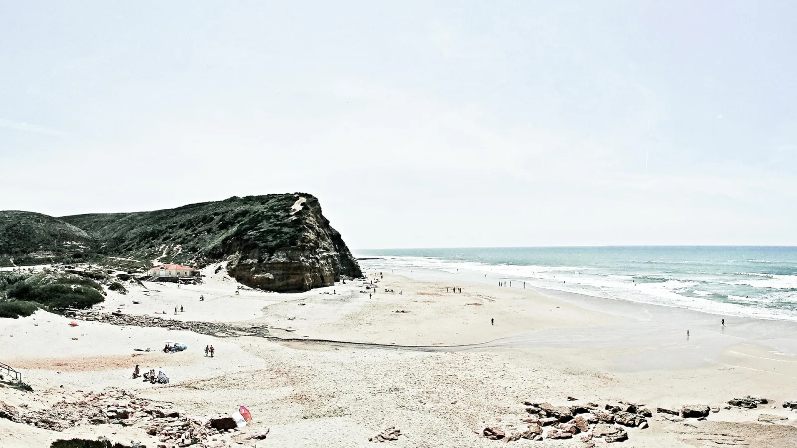 Breiter Sandstrand mit einer steilen, bewachsenen Felsklippe links, einem kleinen weißen Haus mit rotem Dach und verstreuten Badegästen. Rechts rollen Wellen auf das Meer zu, darüber ein hellgrauer Himmel.