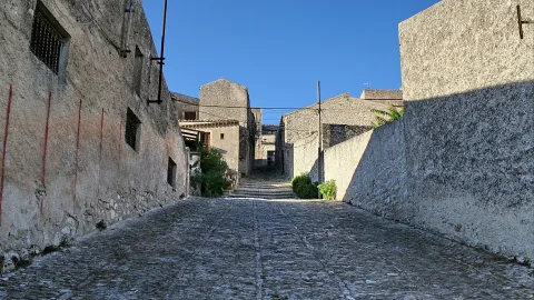 Eine steil ansteigende Kopfsteinpflastergasse zwischen hellen Steinmauern unter klarem blauem Himmel, an deren Ende weitere Häuser und Treppen zu sehen sind.