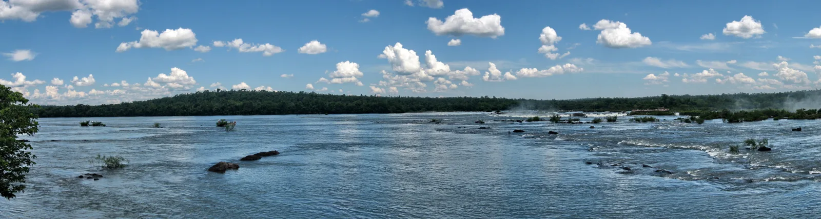 Breiter Fluss mit blaugrauem Wasser, Felsen und Inseln im Vordergrund, dahinter dichter grüner Waldstreifen und weiße Wolken am blauen Himmel, rechts Gischt eines Wasserfalls.