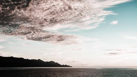 Schichtwolken über einem ruhigen Meer, links ein dunkler Gebirgskamm am Horizont, rechts blassblauer Himmel.
