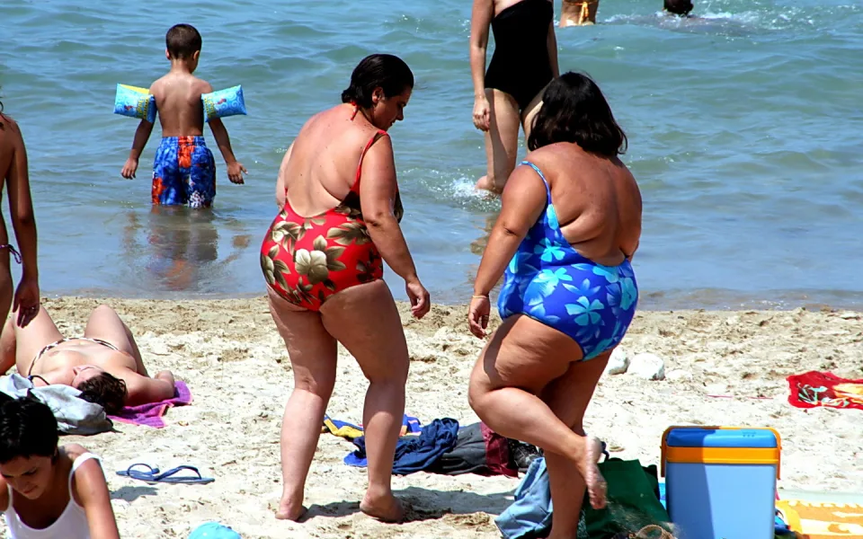 Mehrere Badegäste am Sandstrand: Zwei Frauen in bunten Badeanzügen gehen zum Wasser, im flachen Meer steht ein Kind mit Schwimmflügeln, links liegt eine Person auf einem Handtuch.