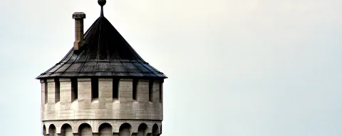 Runder Turm mit spitzem, dunklem Schieferdach, Wetterfahne und Schornstein, unten Rundbögen, rechts heller grauer Himmel