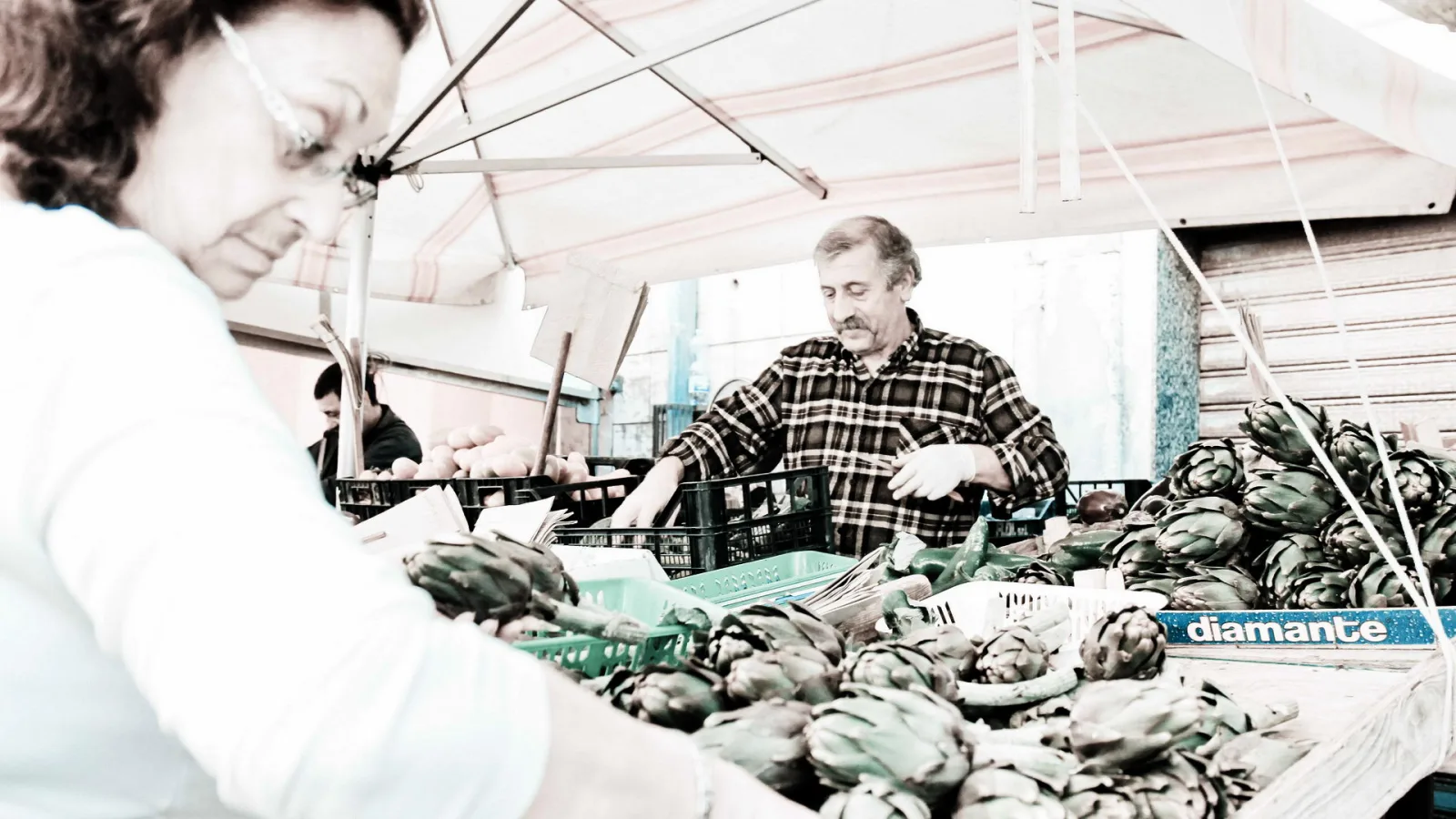 Zwei Marktverkäufer hinter einem Stand mit Artischocken in Kisten, im Vordergrund eine Frau mit Brille, im Hintergrund ein Mann im karierten Hemd unter einer gestreiften Markise.