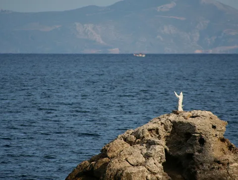 Weiße Jesusfigur mit erhobenen Armen auf einem Felsen am Meer, im Hintergrund Berge und ein kleines Schiff am Horizont.