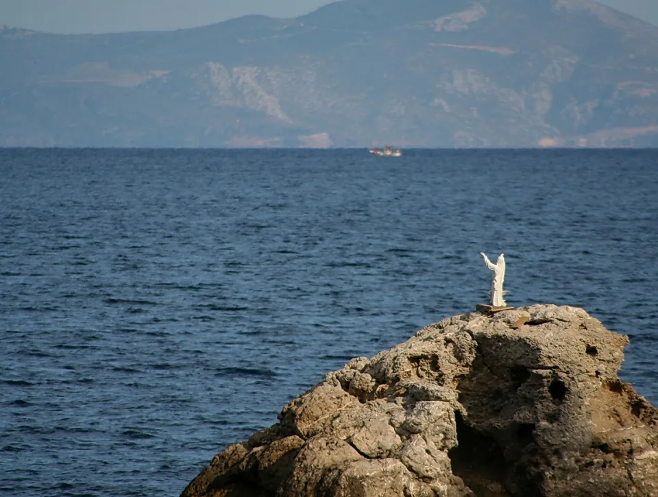 Weiße Jesusfigur mit erhobenen Armen auf einem Felsen am Meer, im Hintergrund Berge und ein kleines Schiff am Horizont.
