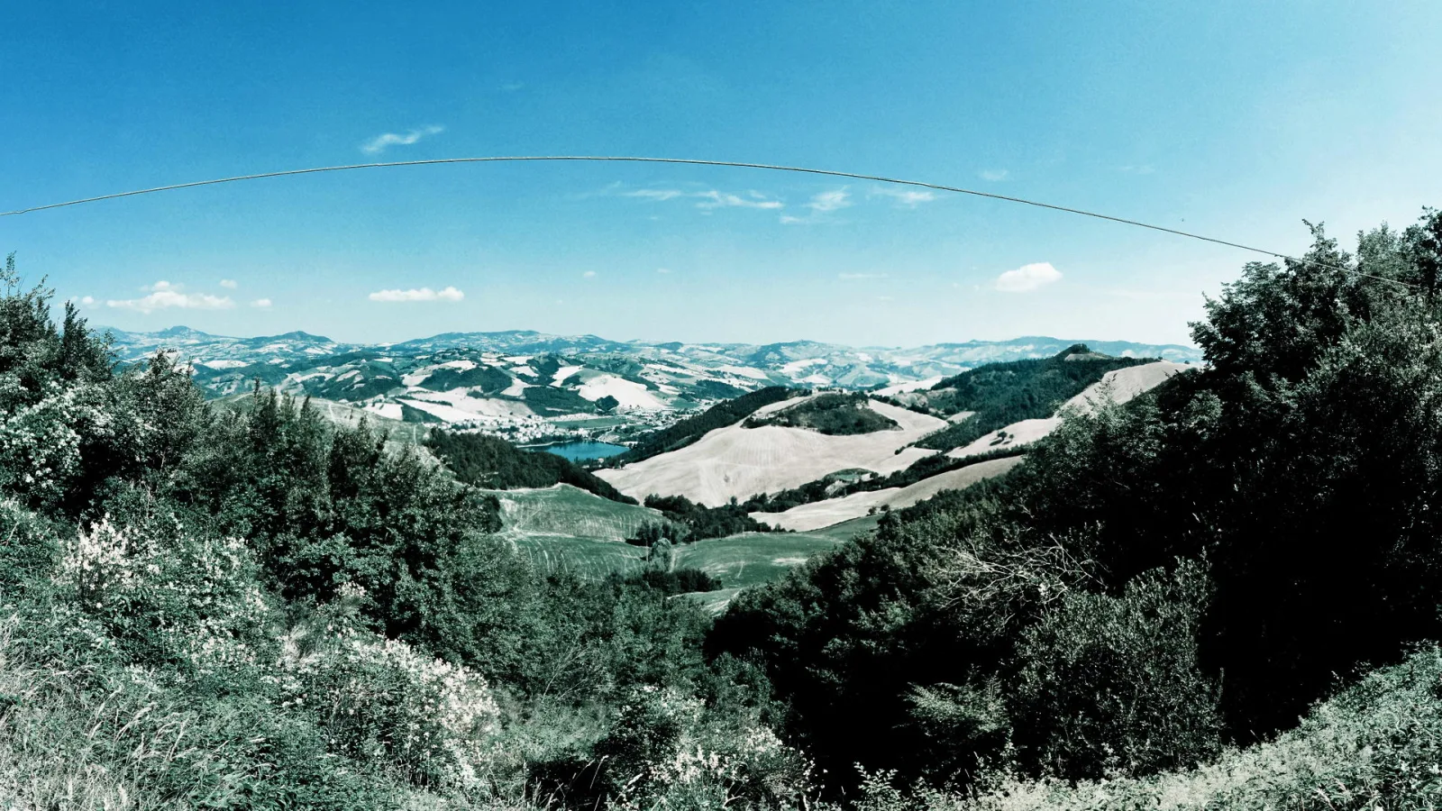 Panoramablick über bewaldete und landwirtschaftlich genutzte Hügel unter blauem Himmel, im Vordergrund dichte Büsche, im Tal ein kleiner See, über dem Bild verläuft eine Stromleitung.