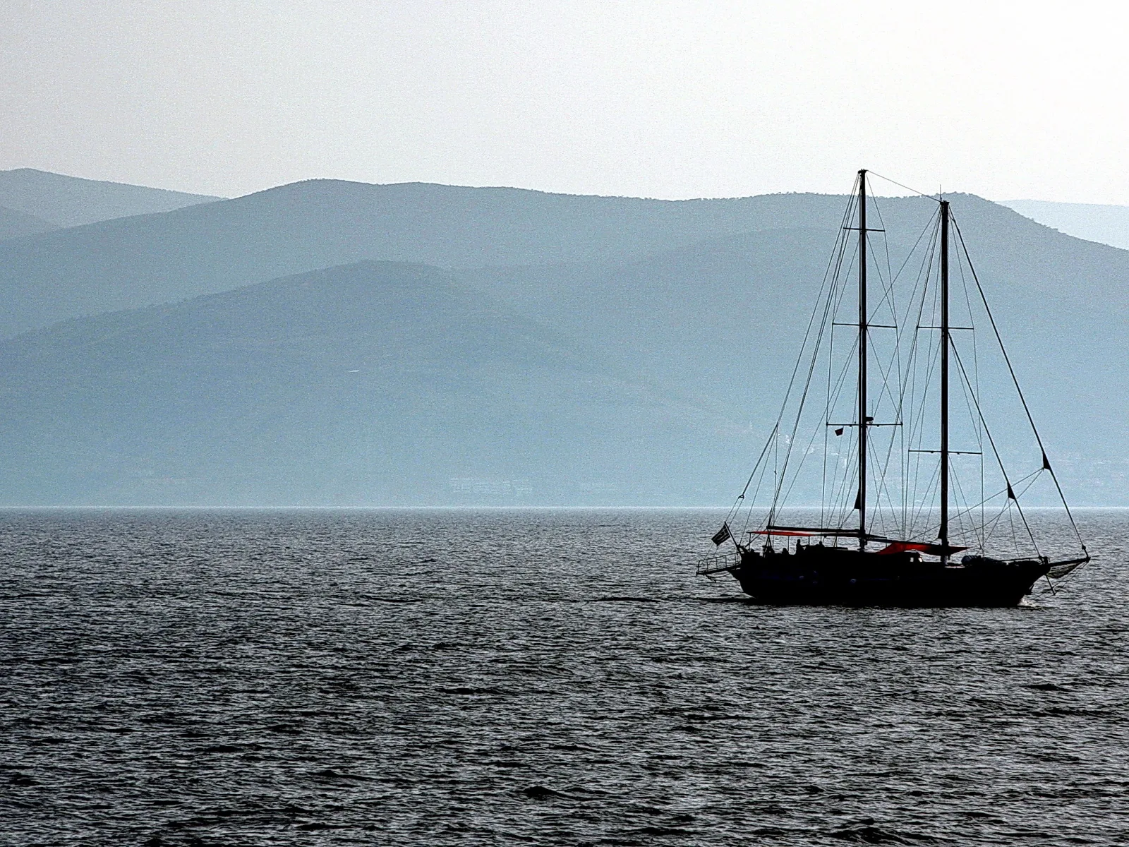 Dunkles Segelschiff mit zwei Masten auf ruhigem Meer, im Hintergrund blaugraue Bergketten unter hellem Dunst.