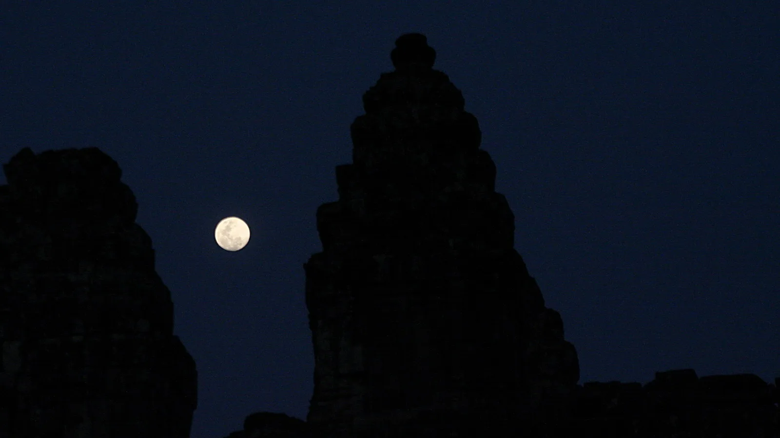 Silhouette eines hohen Tempelturms mit gestuften Spitzen vor dunklem Nachthimmel, links daneben ein Vollmond.