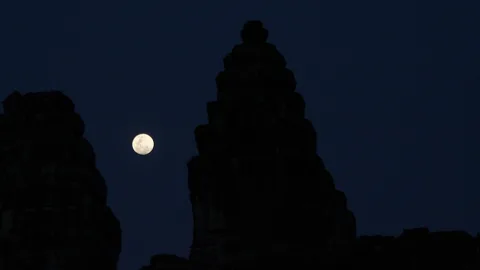 Silhouette eines hohen Tempelturms mit gestuften Spitzen vor dunklem Nachthimmel, links daneben ein Vollmond.