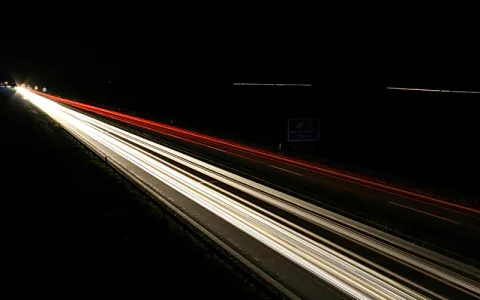 Langzeitbelichtung einer nächtlichen Autobahn: weiße Lichtspuren der Fahrzeuge in Richtung Horizont und rote Rücklichter auf der rechten Fahrbahn, dunkler Himmel, ein blaues Hinweisschild rechts.