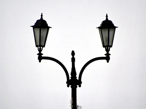 Schwarze, verzierte Doppelstraßenlaterne mit zwei Laternen vor einem hellgrauen, wolkenlosen Himmel.