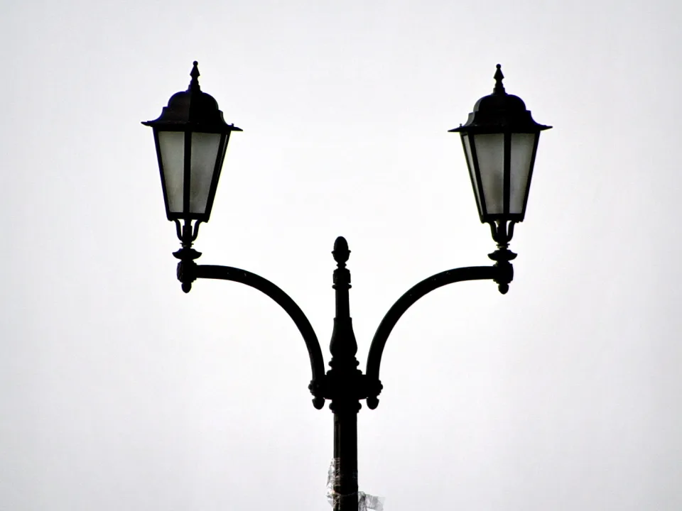 Schwarze, verzierte Doppelstraßenlaterne mit zwei Laternen vor einem hellgrauen, wolkenlosen Himmel.
