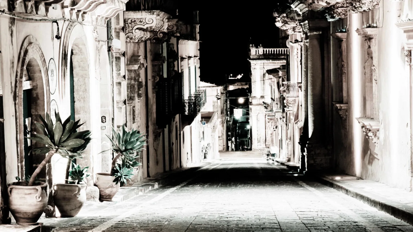Leere, gepflasterte Gasse bei Nacht, gesäumt von barocken Steinhäusern mit verzierten Balkonen und Topfpflanzen im Vordergrund