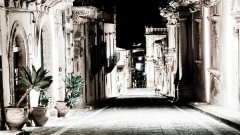 Leere, gepflasterte Gasse bei Nacht, gesäumt von barocken Steinhäusern mit verzierten Balkonen und Topfpflanzen im Vordergrund