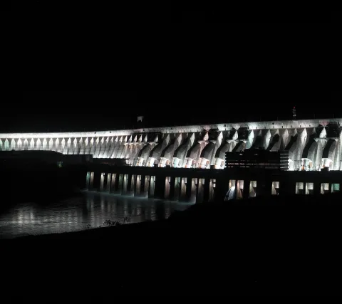 Beleuchtete Staumauer aus Beton bei Nacht, davor ein dunkler Fluss mit Lichtreflexen.