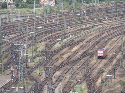 Viele Gleise und Weichen eines Bahngeländes; rechts fährt eine rote Lokomotive durch das Gleisfeld, dazwischen Oberleitungsmasten.