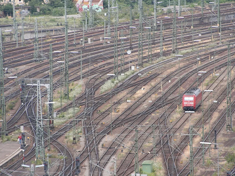 Viele Gleise und Weichen eines Bahngeländes; rechts fährt eine rote Lokomotive durch das Gleisfeld, dazwischen Oberleitungsmasten.