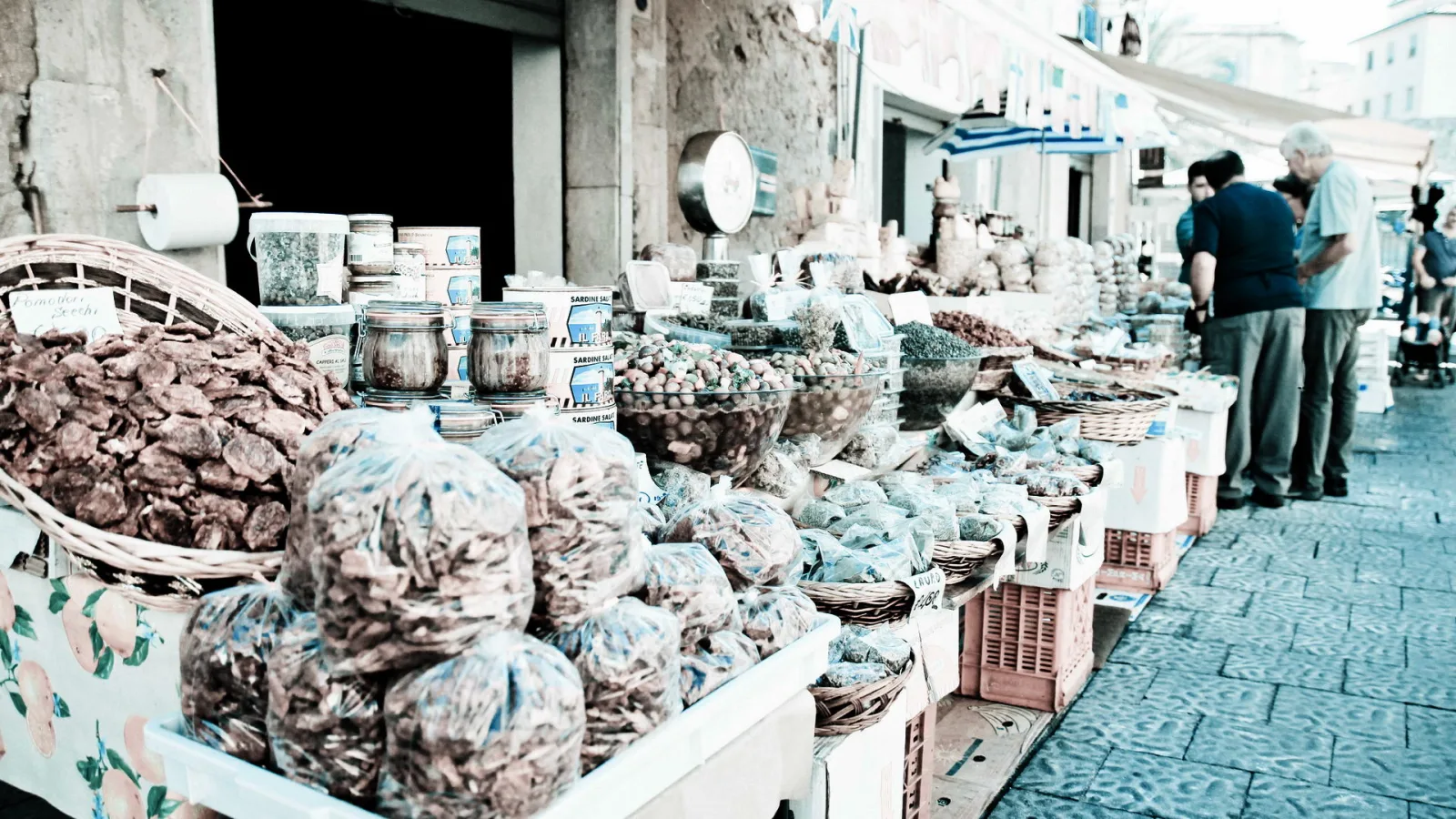 Marktstand mit getrockneten Pilzen oder Früchten in Plastiktüten, Schalen mit Oliven und Gewürzen, daneben zwei ältere Männer beim Einkaufen auf einer gepflasterten Gasse.
