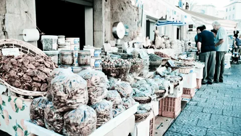 Marktstand mit getrockneten Pilzen oder Früchten in Plastiktüten, Schalen mit Oliven und Gewürzen, daneben zwei ältere Männer beim Einkaufen auf einer gepflasterten Gasse.