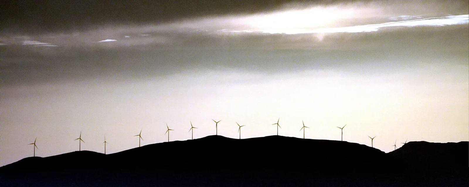 Silhouette einer Hügelkette mit einer Reihe Windkraftanlagen vor einem hellen, bewölkten Abendhimmel