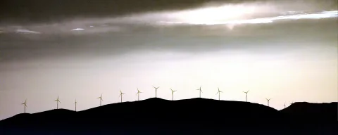 Silhouette einer Hügelkette mit einer Reihe Windkraftanlagen vor einem hellen, bewölkten Abendhimmel