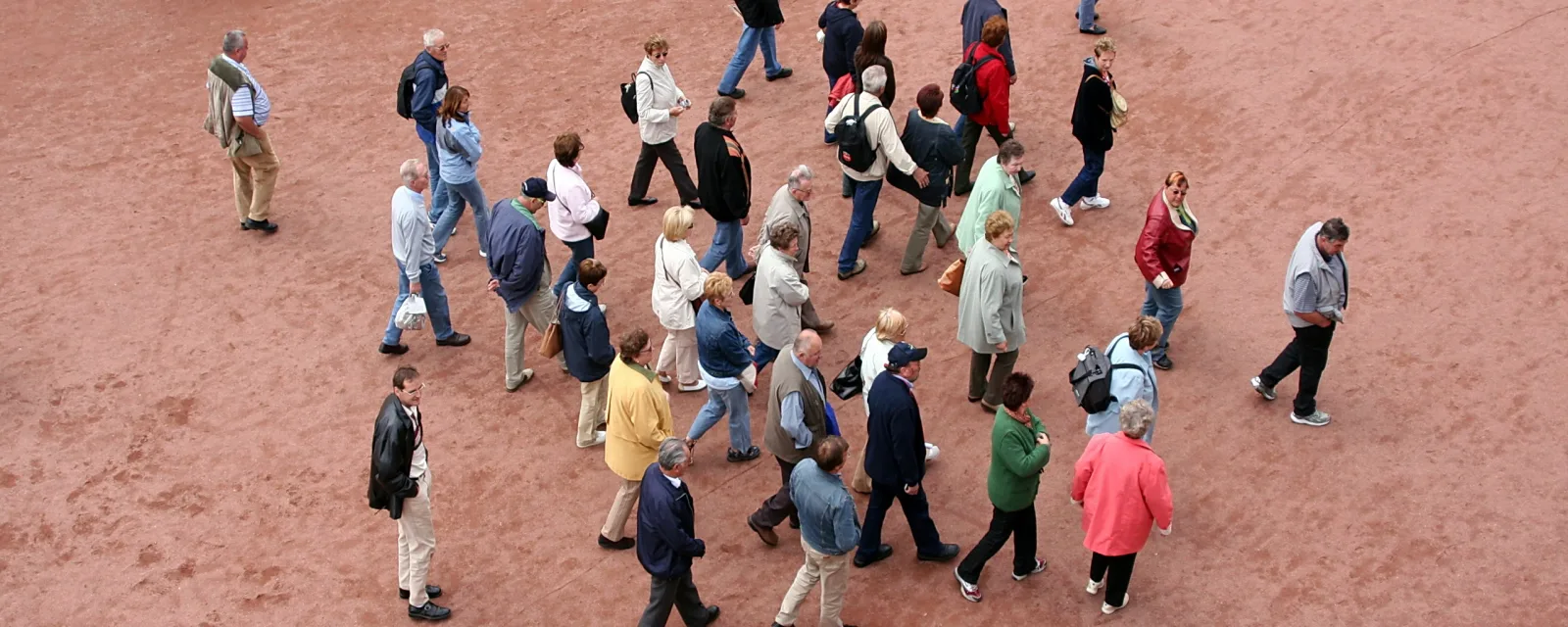 Luftaufnahme von vielen Menschen, die über einen rötlich-sandfarbenen Platz gehen, die meisten in Jacken und hellen Hosen.