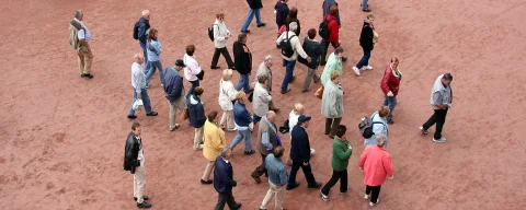Luftaufnahme von vielen Menschen, die über einen rötlich-sandfarbenen Platz gehen, die meisten in Jacken und hellen Hosen.
