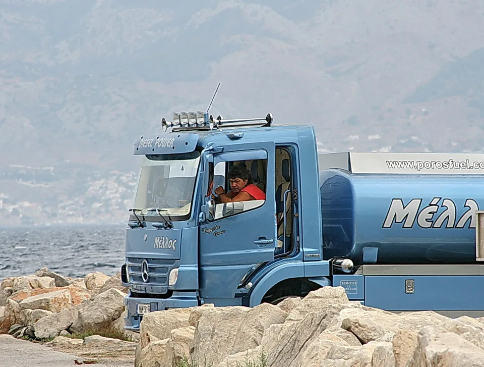 Blauer Tankwagen mit der Aufschrift Μέλλος steht an einer Felsenküste, am Steuer sitzt ein Mann im roten Shirt, dahinter liegen Meer und dunstige Berge.