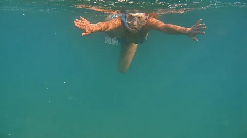 Frau mit Schnorchelmaske schwimmt unter Wasser, beide Arme weit ausgebreitet, blaugrünes Meer